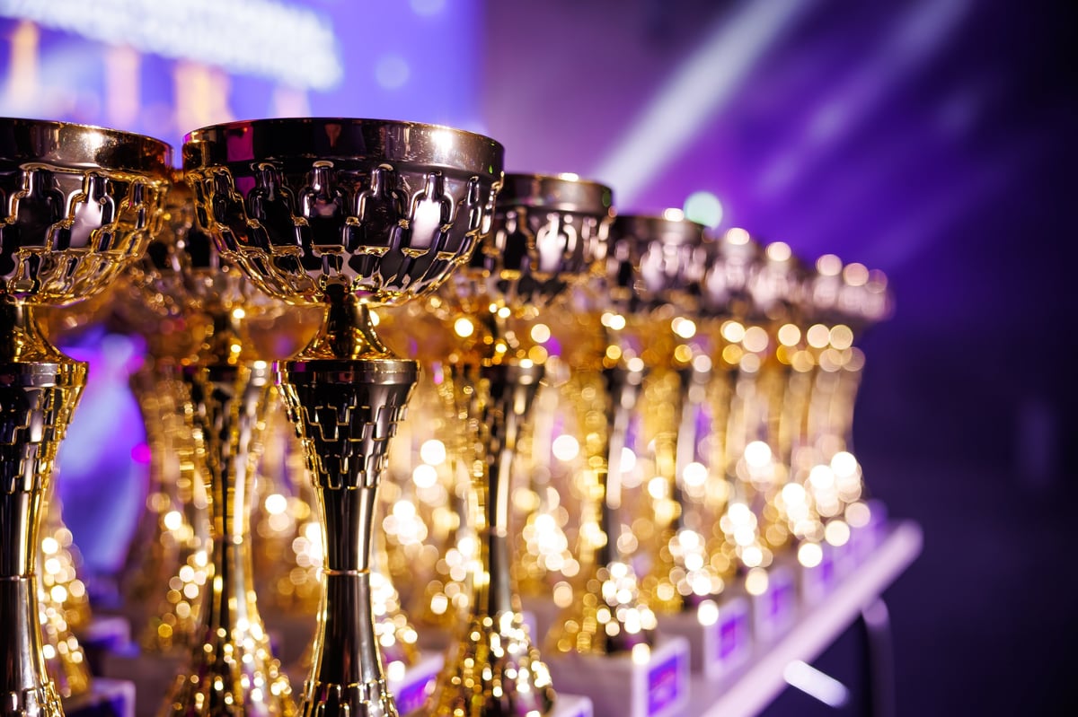 Dozens of ornate gold and silver trophies line a table, lattice bowls and tall stems gleam under purple and white spotlights in an indoor awards ceremony scene.