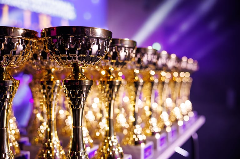 Dozens of ornate gold and silver trophies line a table, lattice bowls and tall stems gleam under purple and white spotlights in an indoor awards ceremony scene.