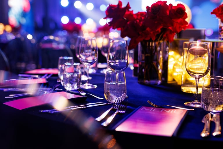 A formally set banquet table with glassware, menus, and floral centerpieces under blue atmospheric lighting.